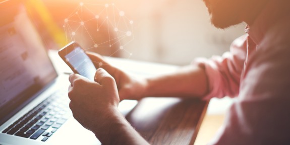 Mann in rosa Hemd sitzt an einem Schreibtisch mit Laptop und hält ein Smartphone in der Hand – symbolisiert mobiles Arbeiten oder digitale Kommunikation im modernen Arbeitsumfeld