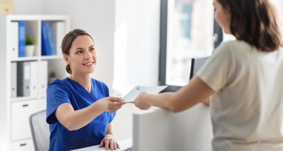 Freundliche medizinische Fachangestellte in blauer Kleidung reicht einer Patientin an der Rezeption ein Dokument – Szene in einer modernen Arztpraxis bei Tageslicht.