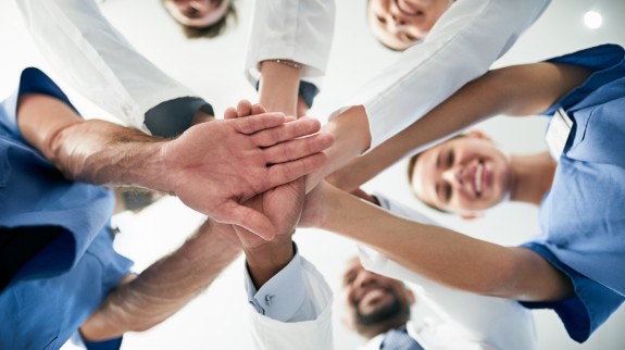 Gruppe von medizinischen Fachkräften steht im Kreis und legt motiviert die Hände übereinander – Symbolbild für Teamgeist, Zusammenarbeit und gemeinsames Engagement im Gesundheitswesen.
